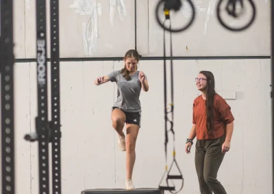 A woman jumps onto a box in a gym while another woman watches and encourages her.