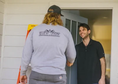 Two men shake hands at the front door; one wears “Elevation” shirt, the other smiles in a black polo.
