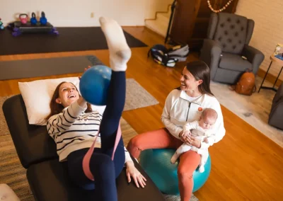 Woman does leg exercise with a ball on a table while another woman holds a baby and smiles nearby.
