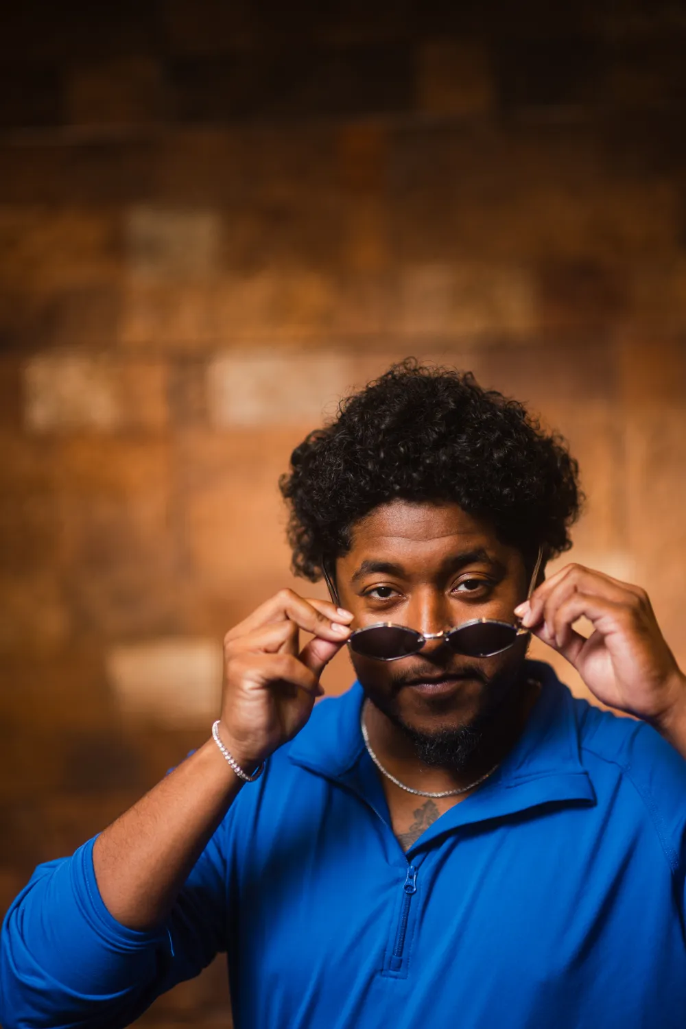 Man in blue shirt lowers sunglasses, looking directly at the camera against a brown, blurred background.