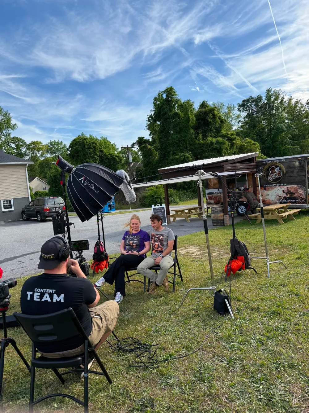 Two people sit for an outdoor video interview as crew films and records with equipment under a blue sky.