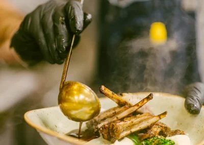 Chef in gloves pours sauce over a plated lamb dish with vegetables and mashed potatoes.
