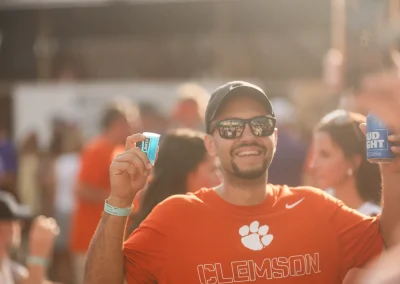 Man in Clemson shirt holding drinks, smiling at an outdoor event with people in the background.