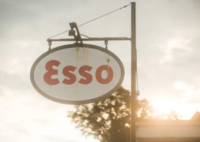 An oval Esso gas station sign hangs outdoors with sunlight and trees in the background.