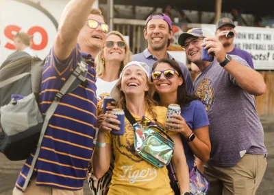 Six adults in casual, colorful clothes smile and take a group selfie outside at an event or festival.