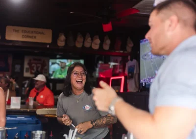 A woman laughs behind a bar while chatting with a man; TVs and decor fill the lively background.
