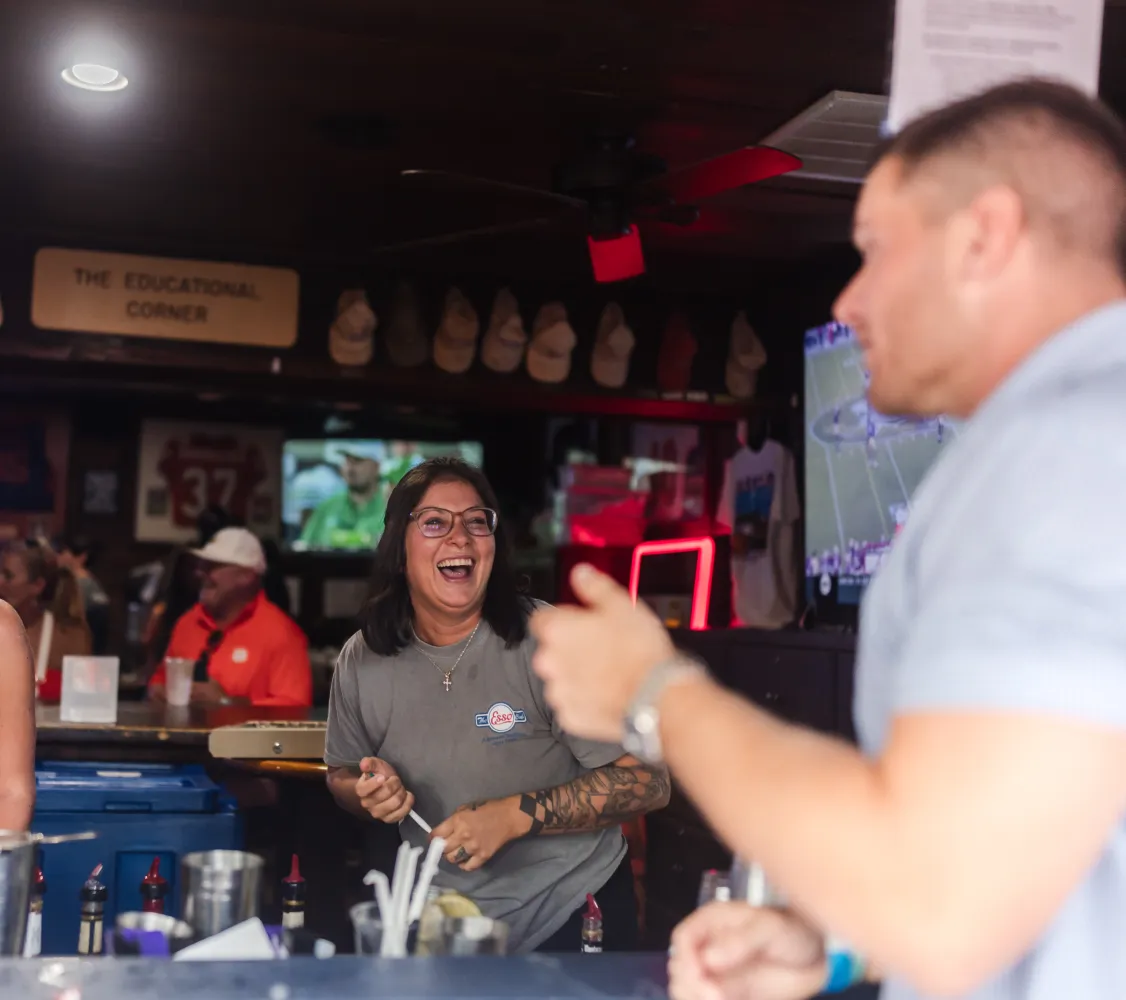 esso-5 A woman laughs behind a bar while chatting with a man; TVs and decor fill the lively background.