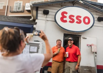 Two men smile for a photo outside an Esso gas station as a woman takes their picture.