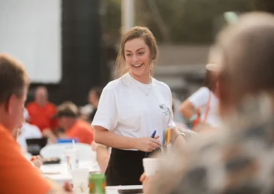 Young woman in a white t-shirt smiling while talking to people seated at tables outdoors.