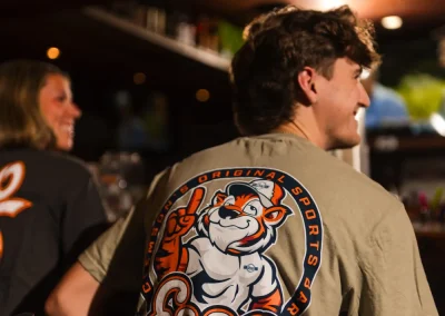 Two people sit at a bar; the man wears a shirt with a cartoon tiger and the text “Esso Club” on the back.