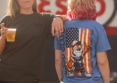 Two women stand together; one holds a drink while the other wears a shirt with a cartoon tiger and USA flag.