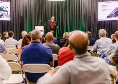 A speaker presents to a seated audience in a conference room with slides displayed on screens.