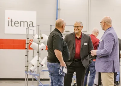 Three men in business attire talk in a factory with the Itema logo and machinery in the background.