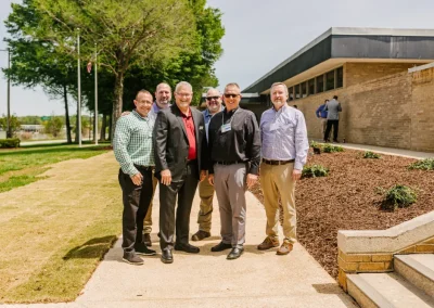 Six men stand smiling outside a tan brick building on a sunny day, near a mulched garden bed.