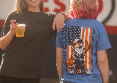 Two women outdoors; one holds a drink, the other wears a cartoon baseball shirt with a US flag design.