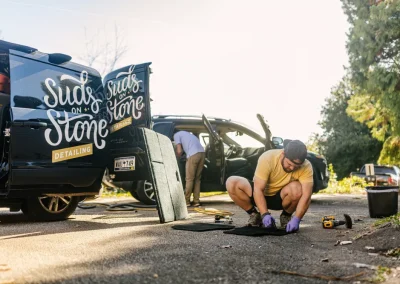 Man in gloves cleans car floor mat outdoors; van with “Suds on Stone Detailing” on the side is nearby.