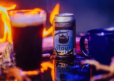 A can of coffee oatmeal stout, a glass, and a mug sit near a campfire with flames in the background.