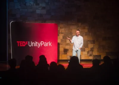 A speaker stands on stage at TEDxUnityPark, addressing an audience in a dimly lit room.