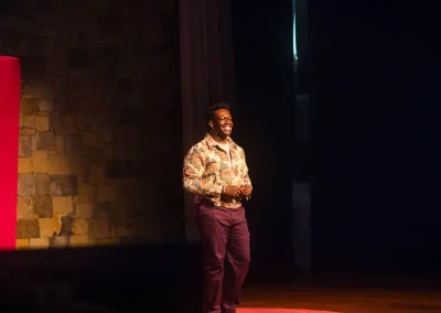 A man stands and smiles on a stage with a bright spotlight, near a red TEDx sign and a stone wall.