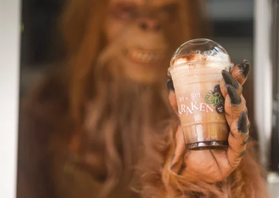 A hairy, ape-like creature holds out a plastic cup of iced coffee labeled “let’s get KRAKEN.”