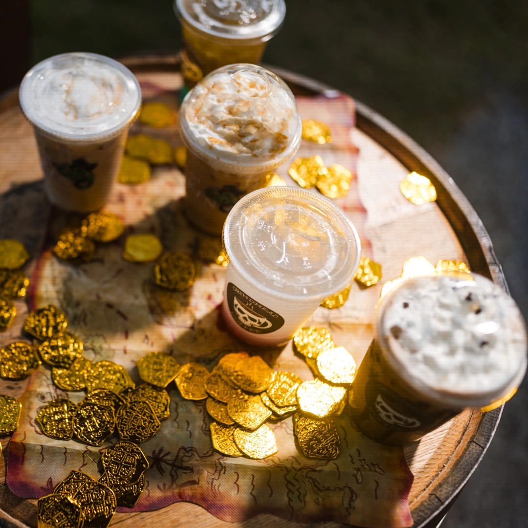 Four iced drinks with whipped cream on a wooden barrel decorated with gold coins and a treasure map.