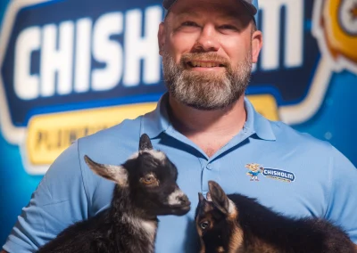 A man wearing a Chisholm company polo and cap holds two small black goats.