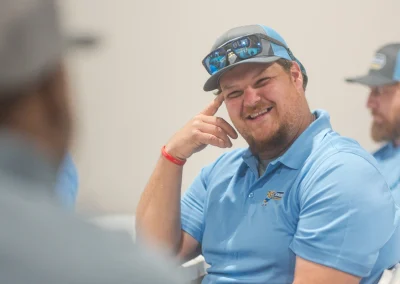 A smiling man in a blue polo shirt and cap sits at a table with others.