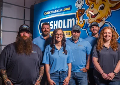Six Chisholm Plumbing and Air employees posing in front of a company-branded wall.