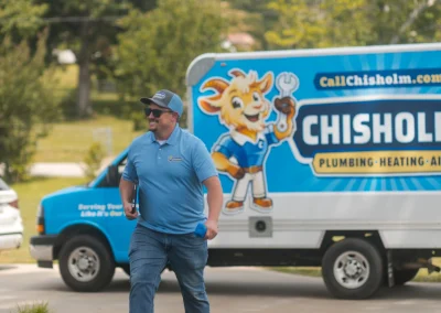 A smiling man in a blue uniform shirt walks in front of a Chisholm Plumbing, Heating, & Air service van.