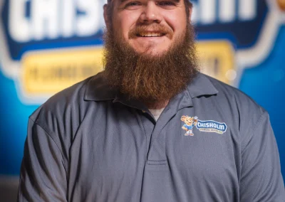 A bearded man wearing a Chisholm Plumbing, Heating, and Air uniform and cap in front of a company logo.