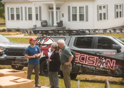 A worker in a blue shirt speaks with two clients in front of a Precision Pest and Wildlife Removal truck.