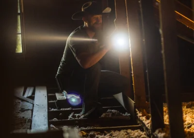 A man in a cowboy hat kneels in an attic, shining a flashlight on wooden rafters.